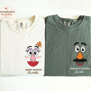 May include: Two crewneck t-shirts, one cream and one sage green, featuring cartoon potato character faces. The cream shirt displays a female-coded face with text "SWEET POTATO Charlotte," while the green shirt shows a male-coded face with text "GROUCHY POTATO Jacobo."