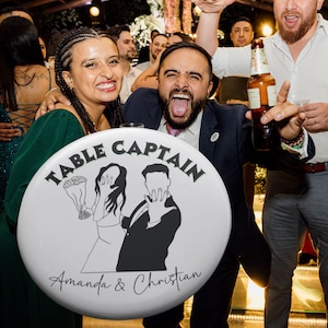 May include: A white button with the words "TABLE CAPTAIN" in a circular design. Below the text is a black and white illustration of a bride and groom. The names "Amanda & Christian" are written below the illustration.
