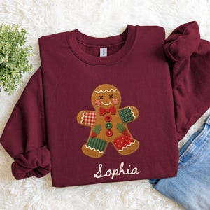 May include: A maroon crewneck sweatshirt featuring a central gingerbread man graphic with patchwork-style patterns in red, green, and white. The gingerbread figure has a red bow tie, three buttons, and the name Sophia printed in white script below.