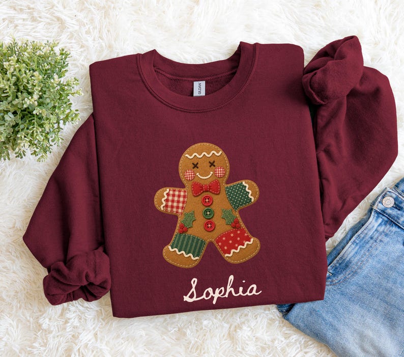 May include: A maroon crewneck sweatshirt featuring a central gingerbread man graphic with patchwork-style patterns in red, green, and white. The gingerbread figure has a red bow tie, three buttons, and the name Sophia printed in white script below.