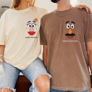 May include: A cream-colored t-shirt with a graphic of a face and the text SWEET POTATO, and a brown t-shirt with a graphic of a face and the text GROUCHY POTATO. Both shirts feature cartoon-style facial features including eyes, noses, and mouths.