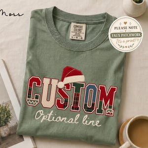 May include: A moss green t-shirt featuring the text CUSTOM in a faux patchwork varsity font with a red and white Santa hat graphic above the letter S. Below the word CUSTOM, the text Optional line is printed in a white script font.