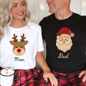 May include: A white t-shirt featuring a brown reindeer graphic with a red nose and the word Mom in green, and a black t-shirt featuring a Santa Claus graphic with a red hat and the word Dad in gold, both paired with red and black plaid pants.