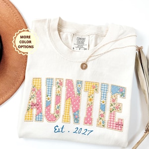 May include: Cream-colored Auntie sweatshirt with patchwork-style lettering in yellow gingham, pink polka dots, blue gingham, and floral patterns. The word "AUNTIE" is embroidered, with "Est. 2027" in blue script below. A gold necklace and a brown hat are also visible.