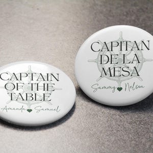 May include: Two white round buttons with nautical-themed designs. One says "CAPTAIN OF THE TABLE" with a ship's wheel illustration and the names "Amanda Samuel". The other says "CAPITAN DE LA MESA" with a ship's wheel and the names "Sammy Nelson".