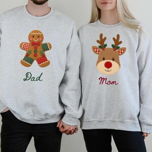 May include: Two light gray crewneck sweatshirts. The left sweatshirt features a brown gingerbread man applique with red and green patterned patches and the green embroidered text "Dad". The right sweatshirt features a brown reindeer applique with red and green accents and the red embroidered text "Mom".