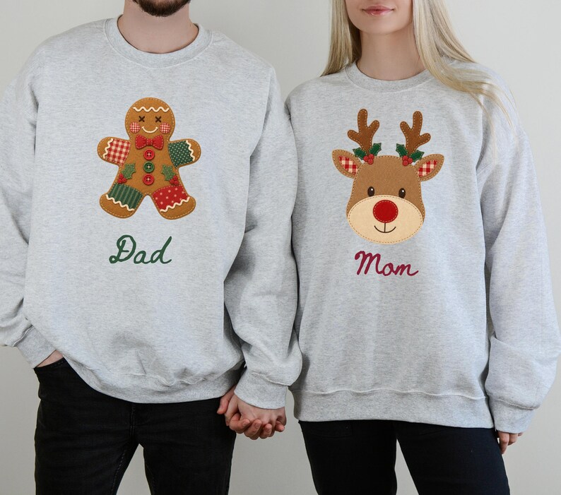 May include: Two light gray crewneck sweatshirts. The left sweatshirt features a brown gingerbread man applique with red and green patterned patches and the green embroidered text "Dad". The right sweatshirt features a brown reindeer applique with red and green accents and the red embroidered text "Mom".