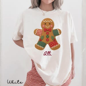 May include: An off-white short-sleeve t-shirt featuring a large gingerbread man graphic with a red and white checkered arm, a green polka dot arm, and a red polka dot leg. The gingerbread man has a red bow tie, three buttons, and the name Alli.