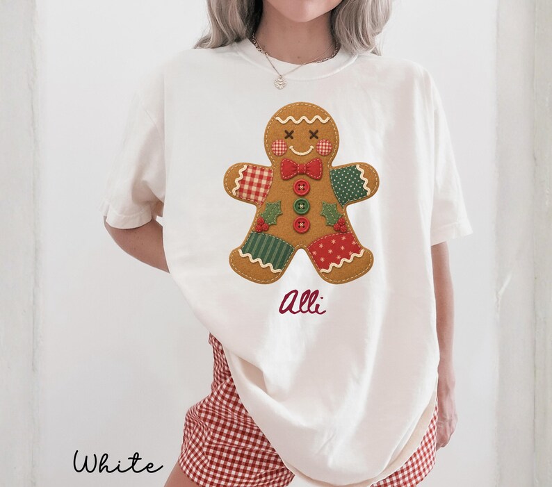 May include: An off-white short-sleeve t-shirt featuring a large gingerbread man graphic with a red and white checkered arm, a green polka dot arm, and a red polka dot leg. The gingerbread man has a red bow tie, three buttons, and the name Alli.