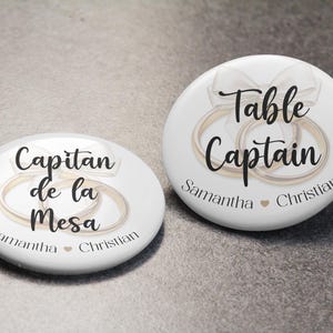 May include: Two white wedding-themed buttons with text and ring graphics. One says "Table Captain" with names "Samantha" and "Christian." The other says "Capitan de la Mesa" with the same names. Both have a bow graphic.