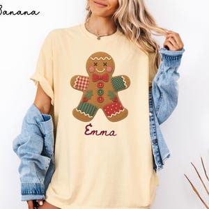 May include: A light yellow t-shirt featuring a gingerbread man graphic with patchwork patterns in red, green, and white. The gingerbread man has a red bow tie and three buttons. The name Emma is printed in red script below the gingerbread man.