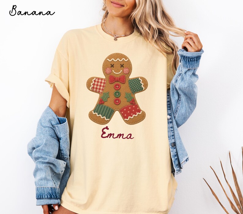 May include: A light yellow t-shirt featuring a gingerbread man graphic with patchwork patterns in red, green, and white. The gingerbread man has a red bow tie and three buttons. The name Emma is printed in red script below the gingerbread man.