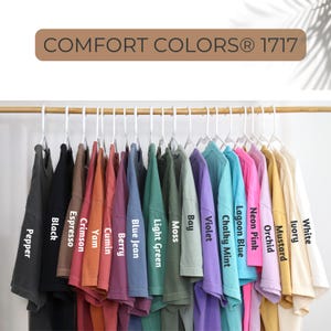 May include: A collection of Comfort Colors 1717 t-shirts hanging on white hangers, featuring a variety of colors labeled as Pepper, Black, Espresso, Crimson, Yam, Cumin, Berry, Blue Jean, Light Green, Moss, Bay, Violet, Chalky Mint, Lagoon Blue, Neon Pink, Orchid, Mustard, Ivory, and White.