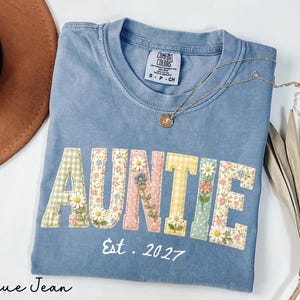 May include: Blue jean colored t-shirt with the word "AUNTIE" in colorful floral and gingham patchwork letters. The text "Est. 2027" is below the word. A gold necklace with a small pendant is on the shirt.