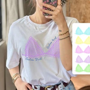 May include: A white short-sleeved t-shirt featuring a purple graphic of a triangle-shaped bralette with a grid pattern and ruffled edges. The text "Custom text" is printed in blue script near the graphic. A color palette shows five options.