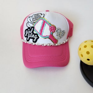May include: Pink and white trucker hat with a white mesh back. The hat has a patch with a pink and blue paddle, a white rose, and the text "girl gang".
