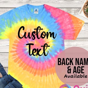 Puede incluir: Una camiseta tie-dye con un patrón de espiral en tonos de azul, amarillo, rosa y naranja. La camiseta tiene el texto "Custom Text" impreso en negro en la parte delantera. El texto "Back Name & Age Available" está impreso en negro sobre un círculo rosa.