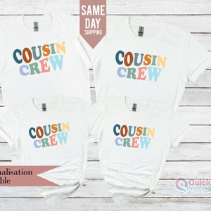 Può includere: Quattro magliette bianche a maniche corte disposte a griglia, ognuna con la scritta "COUSIN CREW" stampata in un carattere ondulato multicolore. Le lettere sono colorate in tonalità di blu, arancione, giallo, verde e viola.
