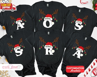 May include: Black t-shirts with a Christmas reindeer design. Each shirt features a reindeer antler design, a Santa hat with a name, and a large initial. The names include Amelia, Olivia, Sophia, and Rowen.