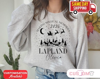 Può includere: Una felpa girocollo grigio chiaro con una stampa grafica nera raffigurante un villaggio invernale con alberi di pino, una luna crescente, stelle e Babbo Natale su una slitta. Il testo recita "MAKING MAGICAL MEMORIES 2026 LAPLAND Olivia" in nero.
