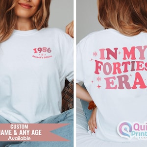 May include: White t-shirt with pink text. The front of the shirt has the year "1986" and "Hannah's Edition". The back of the shirt says "IN MY FORTIES ERA". The bottom of the image says "CUSTOM NAME & ANY AGE Available".