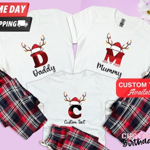 Puede incluir: Un conjunto de tres camisetas blancas con pantalones de pijama de cuadros rojos y negros. Las camisetas presentan una letra de cuadros rojos y negros con una diadema de cuernos de reno y un gorro de Papá Noel. Las letras son "D" para "Papá", "M" para "Mamá" y "C" para "Texto personalizado".