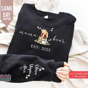 May include: Black sweatshirt featuring a Winnie the Pooh graphic with the words "mama bear" and "EST: 2025". The design includes bees and a honey pot. The sleeve displays names and bee graphics. A "Customisation Available" tag is visible.