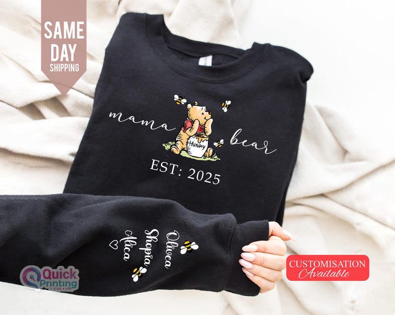 May include: Black sweatshirt featuring a Winnie the Pooh graphic with the words "mama bear" and "EST: 2025". The design includes bees and a honey pot. The sleeve displays names and bee graphics. A "Customisation Available" tag is visible.