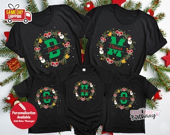 May include: Black family Christmas t-shirts with personalized wreath designs. Each shirt features a letter and name, surrounded by a wreath with bows, bells, and snowmen. The text "Dada", "Mama", "Caleb", "Henry", and "Olivia" are visible.