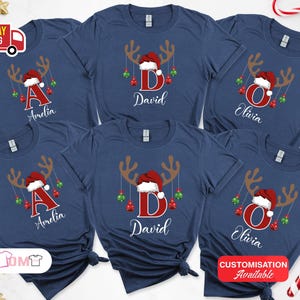 Pode incluir: T-shirts azul marinho com um design de rena de Natal. Cada camisa apresenta uma letra e um nome em branco, com um gorro de Papai Noel e enfeites pendurados. Personaliza&ccedil;&atilde;o dispon&iacute;vel.