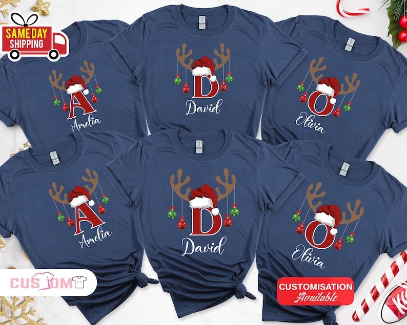 Pode incluir: T-shirts azul marinho com um design de rena de Natal. Cada camisa apresenta uma letra e um nome em branco, com um gorro de Papai Noel e enfeites pendurados. Personaliza&ccedil;&atilde;o dispon&iacute;vel.