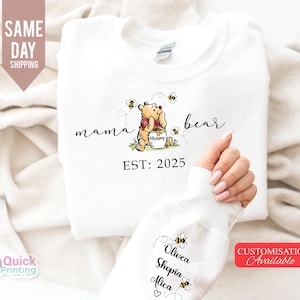 May include: White sweatshirt featuring a Winnie the Pooh design with the words "mama bear" and "EST: 2025". The design includes a cartoon bear, bees, and a honey pot. The sleeve displays names and bee graphics.