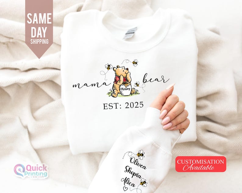May include: White sweatshirt featuring a Winnie the Pooh design with the words "mama bear" and "EST: 2025". The design includes a cartoon bear, bees, and a honey pot. The sleeve displays names and bee graphics.