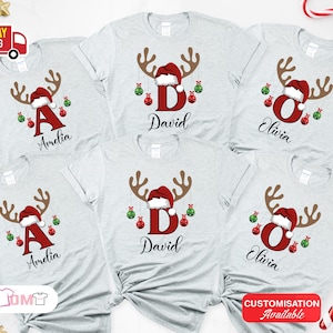Pode incluir: T-shirts cinzentas claras com um design de rena de Natal. Cada camisa apresenta uma letra, um nome, um gorro de Papai Noel, chifres e enfeites pendurados. As camisas s&atilde;o personalizadas com nomes como Amelia, David e Olivia.