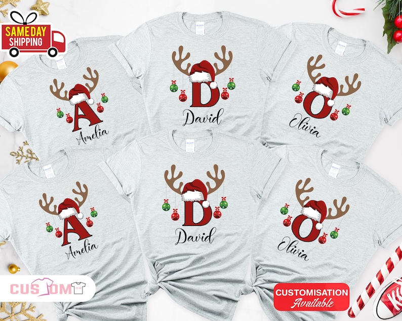 Pode incluir: T-shirts cinzentas claras com um design de rena de Natal. Cada camisa apresenta uma letra, um nome, um gorro de Papai Noel, chifres e enfeites pendurados. As camisas s&atilde;o personalizadas com nomes como Amelia, David e Olivia.