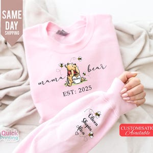 May include: Pink sweatshirt featuring a Winnie the Pooh design with the words "mama bear" and "EST: 2025". The sleeve displays names and bee graphics. The image also includes text that says "Same Day Shipping" and "Customisation Available".