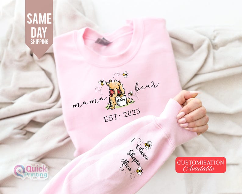 May include: Pink sweatshirt featuring a Winnie the Pooh design with the words "mama bear" and "EST: 2025". The sleeve displays names and bee graphics. The image also includes text that says "Same Day Shipping" and "Customisation Available".