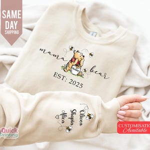 May include: Beige sweatshirt with the text "mama bear" and a Winnie the Pooh graphic. The sweatshirt has the text "EST: 2025" and names on the sleeve. The design includes bees. Customization is available.