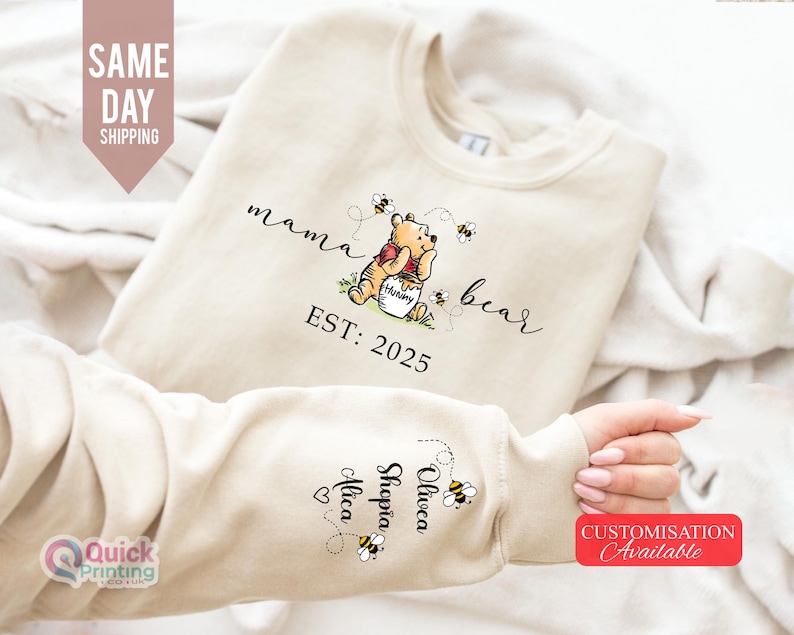 May include: Beige sweatshirt with the text "mama bear" and a Winnie the Pooh graphic. The sweatshirt has the text "EST: 2025" and names on the sleeve. The design includes bees. Customization is available.
