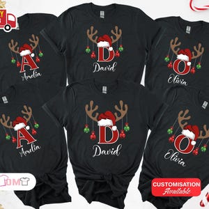 Pode incluir: T-shirts pretas com tema natal&iacute;cio. Cada t-shirt apresenta um design de chifres de rena com um gorro de Pai Natal, enfeites e uma letra ou nome. Os nomes "Amelia", "David" e "Olivia" s&atilde;o vis&iacute;veis.