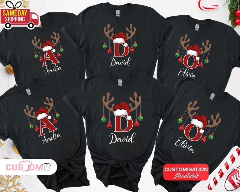 Pode incluir: T-shirts pretas com tema natal&iacute;cio. Cada t-shirt apresenta um design de chifres de rena com um gorro de Pai Natal, enfeites e uma letra ou nome. Os nomes "Amelia", "David" e "Olivia" s&atilde;o vis&iacute;veis.