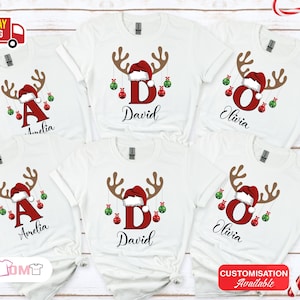 Pode incluir: Camisetas brancas com um design de rena de Natal. Cada camisa apresenta uma letra, um nome e enfeites festivos. As hastes da rena s&atilde;o marrons, o gorro de Papai Noel &eacute; vermelho e os enfeites s&atilde;o vermelhos e verdes.