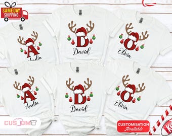May include: White t-shirts with a Christmas reindeer design. Each shirt features a letter, name, and festive ornaments. The reindeer antlers are brown, the Santa hat is red, and the ornaments are red and green.