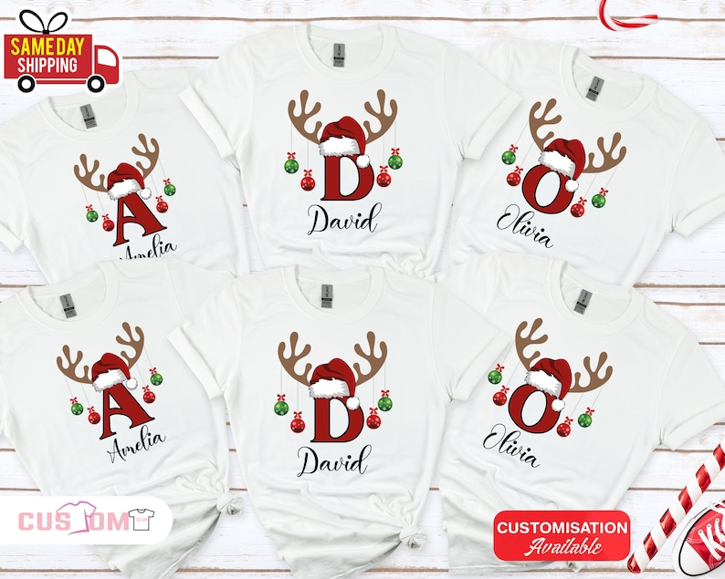 Pode incluir: Camisetas brancas com um design de rena de Natal. Cada camisa apresenta uma letra, um nome e enfeites festivos. As hastes da rena s&atilde;o marrons, o gorro de Papai Noel &eacute; vermelho e os enfeites s&atilde;o vermelhos e verdes.