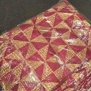 Op de afbeelding: Een gevouwen textiel met een geometrisch patroon van afwisselende bordeauxrode en bruinachtige driehoeken. De stof is verpakt in doorzichtig plastic, wat suggereert dat het nieuw en ongebruikt is. Het ontwerp is ingewikkeld en gedetailleerd.