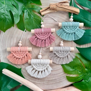 Könnte beinhalten: Fünf Makramee-Wandbehänge mit Holzstäben und Quasten. Die Behänge sind in verschiedenen Farben, darunter rosa, grün, beige und weiß.