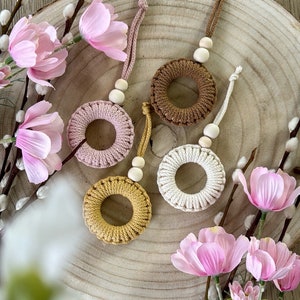 Könnte beinhalten: Vier gehäkelte Ringe in Braun, Rosa, Weiß und Gelb mit Holzperlen, die an jedem Ring hängen. Die Ringe werden auf einer Holzoberfläche mit rosa Blumen präsentiert.