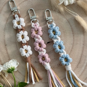 Könnte beinhalten: Drei gehäkelte Blumen-Schlüsselanhänger mit Quasten. Die Anhänger sind in den Farben Weiß, Braun, Rosa und Blau gehalten. Die Schlüsselanhänger sind an silbernen Schlüsselringen befestigt.