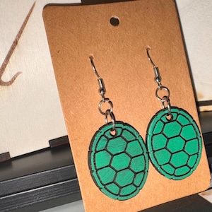Puede incluir: Un par de pendientes de caparazón de tortuga verdes y negros. Los pendientes están hechos de madera y presentan un patrón geométrico detallado. Cuelgan de ganchos para la oreja de color plateado.