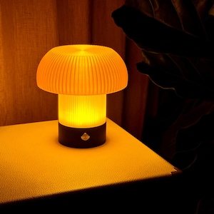 May include: A mushroom-shaped lamp with a ribbed, orange lampshade and a black base. The lamp emits a warm, yellow glow. The lamp is on a yellow surface.
